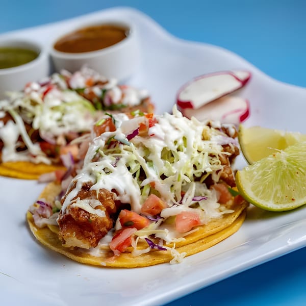 Two Baja California Tacos with fried fish, cabbage, pico de gallo, and crema, served with lime and sauces.