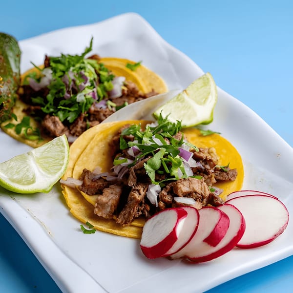 Two Baja California Tacos with meat, cilantro, onions, lime wedges, and sliced radishes on a white plate.